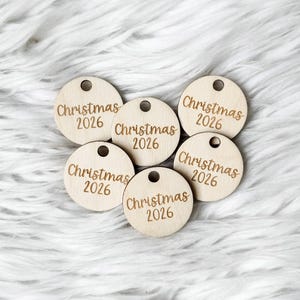 May include: Five circular light-colored wooden tags, each featuring a small hole at the top for hanging. The text "Christmas 2026" is engraved in a cursive font on the center of each wooden disc, creating a uniform set of holiday ornaments.
