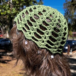 May include: A green crocheted hat with an open weave design. The hat is adorned with small charms, including butterflies and stars. The hat is worn by a person with brown hair.