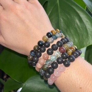 May include: A stack of six beaded bracelets on a person's wrist. The bracelets are made of various stones, including black lava rock, tiger's eye, rose quartz, and green aventurine.