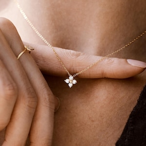 May include: A delicate gold chain necklace featuring a small, four-petalled flower pendant set with tiny clear stones. The necklace is worn around the neck, and a thin gold ring with a small accent is visible on the finger near the pendant.