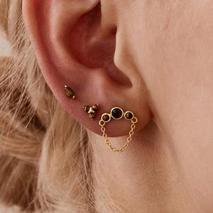 May include: Close-up of an ear adorned with three gold earrings. The earrings include a small stud, a cluster of three dark stones, and a chain-linked design with four dark stones. The ear is set against a blurred background.