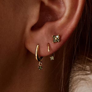 May include: An ear with multiple gold earrings, including small hoops, studs, and dangling designs. Each earring features small green gemstones. The jewellery is set against the backdrop of a person's skin and hair, with a focus on the ear.
