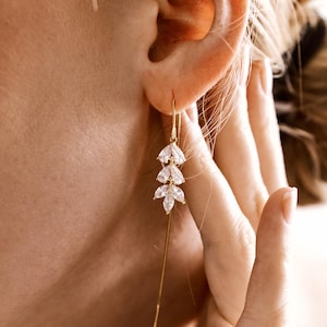 May include: A gold-toned earring with a delicate design featuring three leaf-shaped clusters of clear crystals. The earring hangs from a thin gold chain, creating an elegant and minimalist look. The earring is worn on the ear of a person.