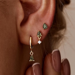 Könnte beinhalten: Nahaufnahme eines Ohrs, das mit drei verschiedenen Arten von goldenen Ohrringen geschmückt ist. Die Ohrringe haben grüne tropfenförmige Edelsteine und kleine weiße Diamanten. Das Ohr befindet sich neben einer Hand mit manikürten Nägeln.