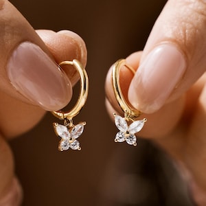 Könnte beinhalten: Goldene Creolen mit einem baumelnden Schmetterlingsanhänger, der mit klaren Zirkonia-Steinen besetzt ist.