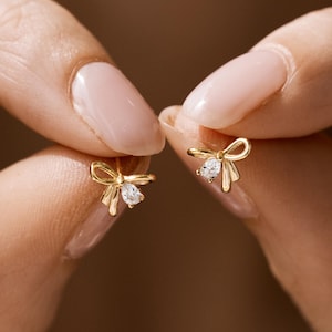 May include: A pair of gold stud earrings shaped like delicate bows. Each earring features a polished gold finish and a single, small, clear, pear-shaped gemstone set at the center of the bow design, adding a subtle sparkle to the metallic structure.