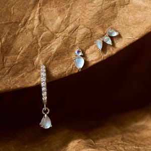 Peut inclure: Trois paires de boucles d'oreilles sont présentées. L'une est un petit cerceau avec des pierres transparentes et une pierre bleue pâle en forme de goutte. Une autre est une puce avec une petite pierre ronde violette et une pierre bleue pâle en forme de goutte. La troisième paire a trois pierres bleues pâles en forme de goutte.