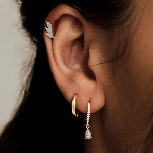 May include: An ear featuring gold earrings. There's a small, curved hoop, a hoop with tiny, clear stones, and a helix earring with three light blue, leaf-shaped stones. The jewellery is set against a blurred background.
