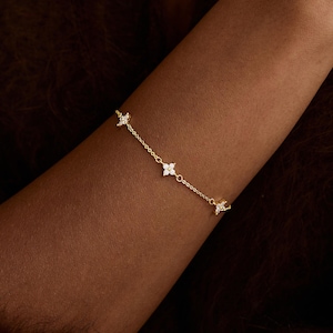 May include: A fine gold bracelet with four star-shaped charms, each set with clear gemstones. The bracelet is displayed on an arm, the gold chain and charms reflecting light. The background is a blurred, dark brown.