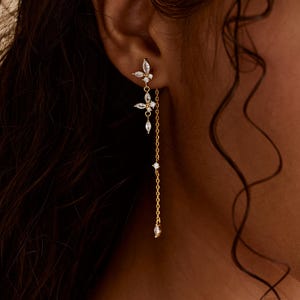 May include: A gold-colored earring with a butterfly design and dangling chain. The earring features clear, sparkling gemstones. The butterfly design is at the ear, with a chain of gemstones and a single dangling gemstone.