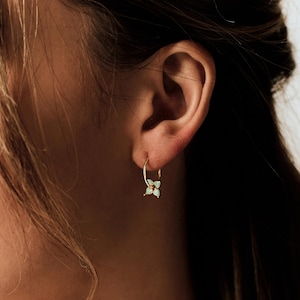 May include: A gold hoop earring featuring a small, four-petaled flower charm with light blue, teardrop-shaped stones. The earring is worn in the earlobe, showcasing a thin gold band and a delicate floral design with a central gold accent.