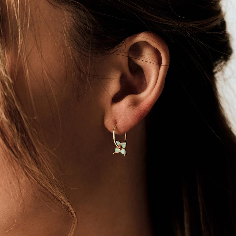 May include: A gold hoop earring featuring a small, four-petaled flower charm with light blue, teardrop-shaped stones. The earring is worn in the earlobe, showcasing a thin gold band and a delicate floral design with a central gold accent.