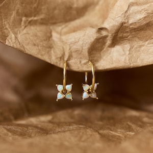 May include: A pair of gold hoop earrings, each featuring a small charm shaped like a four-petaled flower. The petals are made of iridescent white opal stones that display subtle flashes of blue and green color against the polished gold metal.