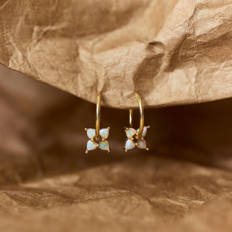 May include: A pair of gold hoop earrings, each featuring a small charm shaped like a four-petaled flower. The petals are made of iridescent white opal stones that display subtle flashes of blue and green color against the polished gold metal.