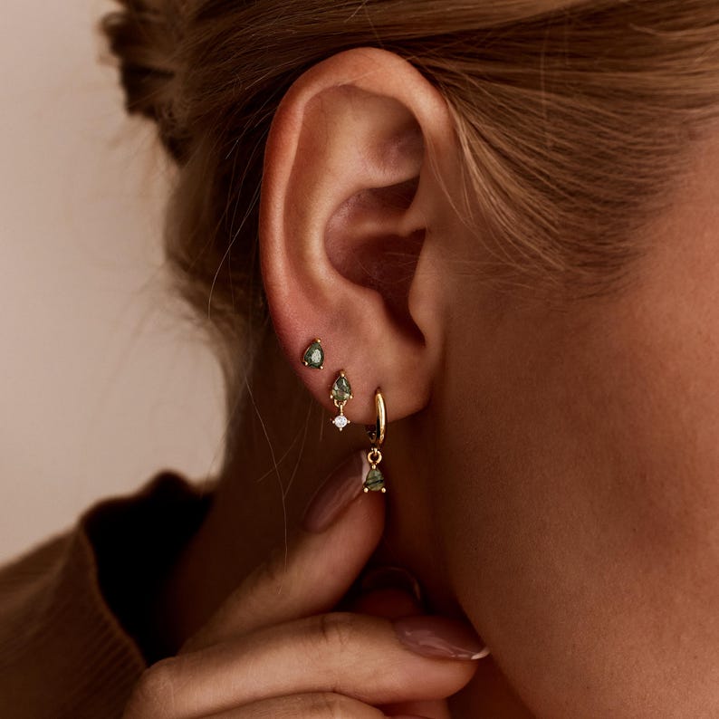 May include: A set of three gold earrings worn in a single ear. The top two are small studs with pear-shaped green stones. The third is a gold hoop earring featuring a dangling pear-shaped green stone and a small clear round stone accent.