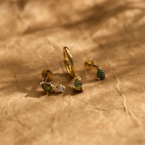 May include: Three gold-toned earrings featuring green pear-shaped stones. One earring is a small hoop with a dangling green stone, while the other two are flat-back studs, one with a single green stone and one with a green stone and a clear crystal.