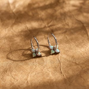 May include: A pair of silver hoop earrings, each featuring a cluster of four small, iridescent, teardrop-shaped opal stones arranged in a flower-like pattern at the base of the hoop. The metal has a polished finish and a circular wire design.