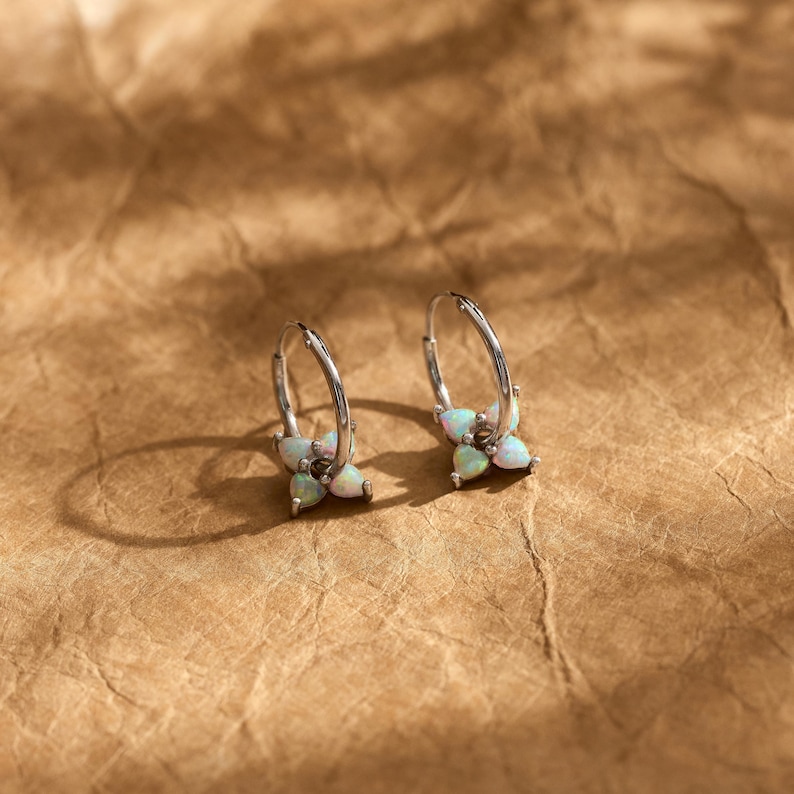 May include: A pair of silver hoop earrings, each featuring a cluster of four small, iridescent, teardrop-shaped opal stones arranged in a flower-like pattern at the base of the hoop. The metal has a polished finish and a circular wire design.