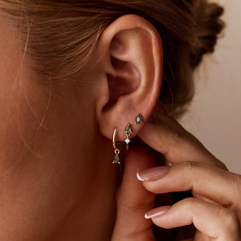 May include: Three gold earrings worn in a single ear. The top earring is a small teardrop-shaped green stone stud. The middle earring is a teardrop-shaped green stone charm hanging from a small clear stone stud. The bottom is a gold hoop with a teardrop-shaped green stone charm.