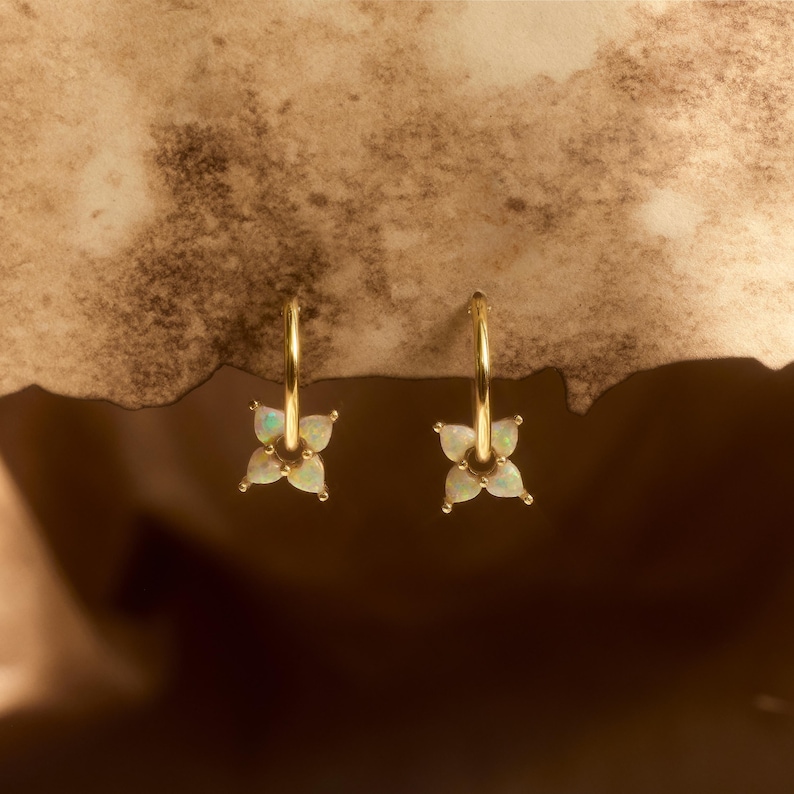 May include: A pair of gold hoop earrings, each featuring a small, four-petaled flower charm. The petals are crafted from iridescent, white opal-like stones that display subtle flashes of color, suspended from a thin, polished gold-tone metal hoop.