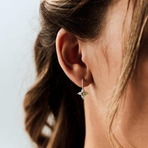 May include: A silver drop earring featuring a small, four-petaled flower design with a central iridescent opal stone. The earring hangs from a thin silver wire hook, resting against the earlobe with a delicate and reflective metallic finish.