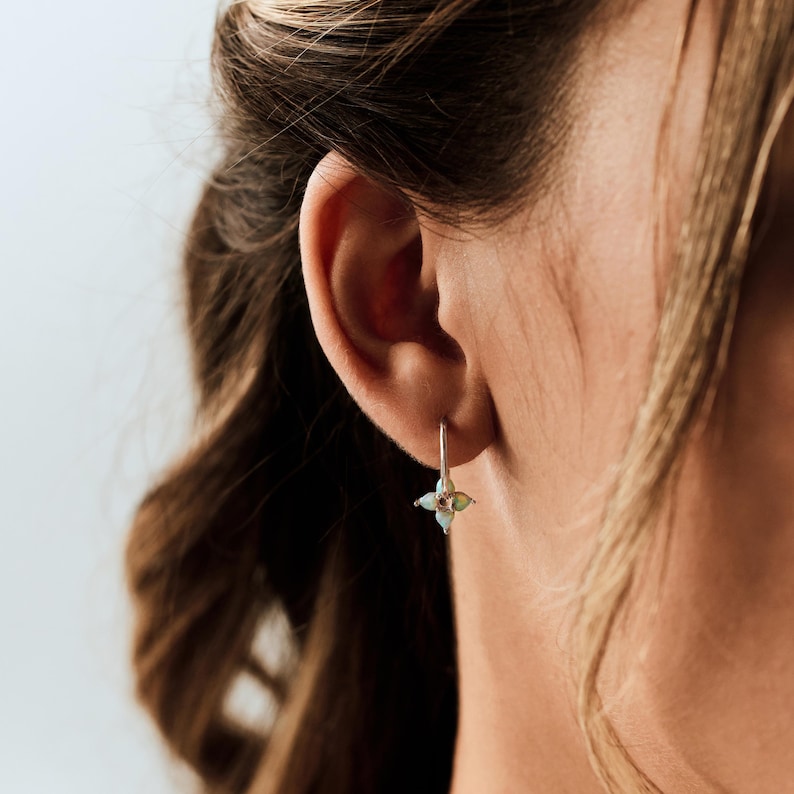 May include: A silver drop earring featuring a small, four-petaled flower design with a central iridescent opal stone. The earring hangs from a thin silver wire hook, resting against the earlobe with a delicate and reflective metallic finish.