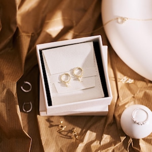 May include: A pair of gold hoop earrings with small clear crystal charms displayed in a white square jewelry box. Additional silver hoop earrings, a gold chain necklace, and a silver ring with small clear stones are arranged nearby.