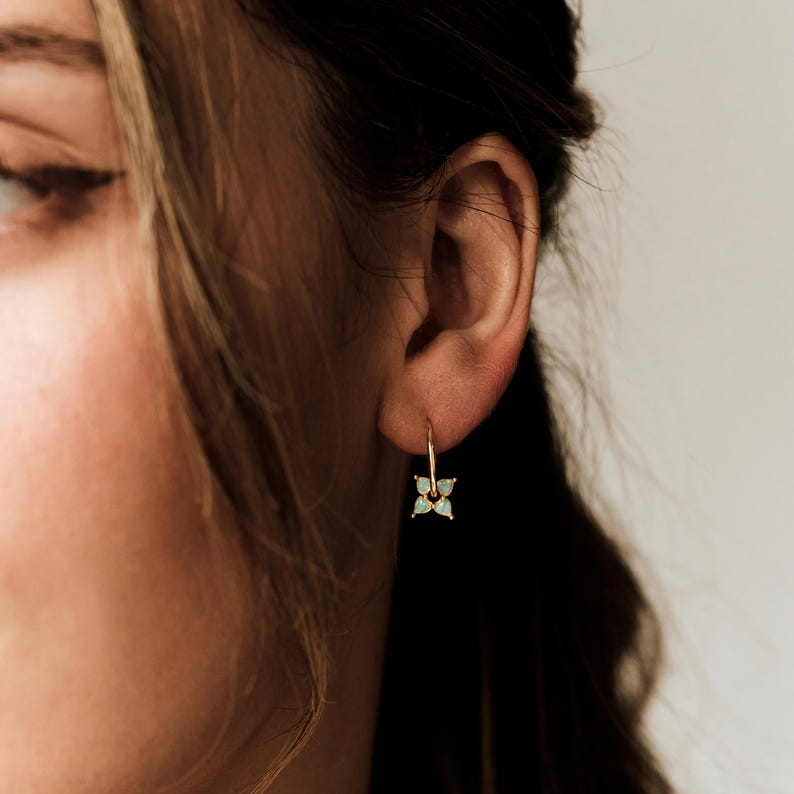May include: A gold hoop earring featuring a small, four-petaled flower charm. The flower is composed of four light blue, teardrop-shaped stones arranged in a symmetrical clover pattern, suspended from a thin, circular gold wire hoop.