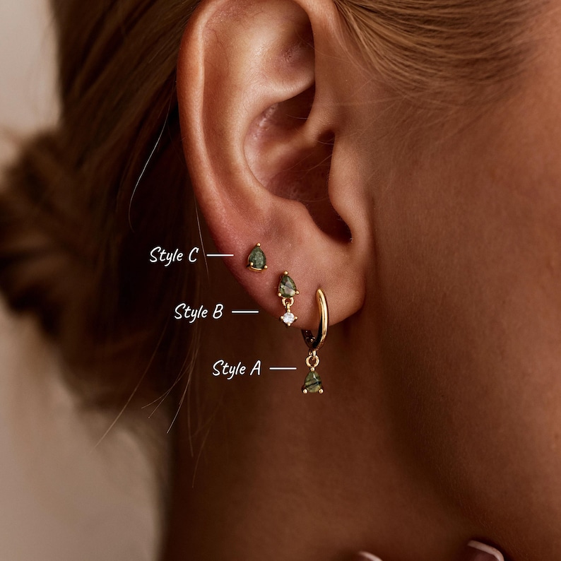 May include: Three gold-toned earrings featuring green pear-shaped stones. Style C is a stud, Style B is a drop earring with a small clear stone, and Style A is a hoop with a dangling green stone. The text labels Style A, Style B, and Style C are visible.