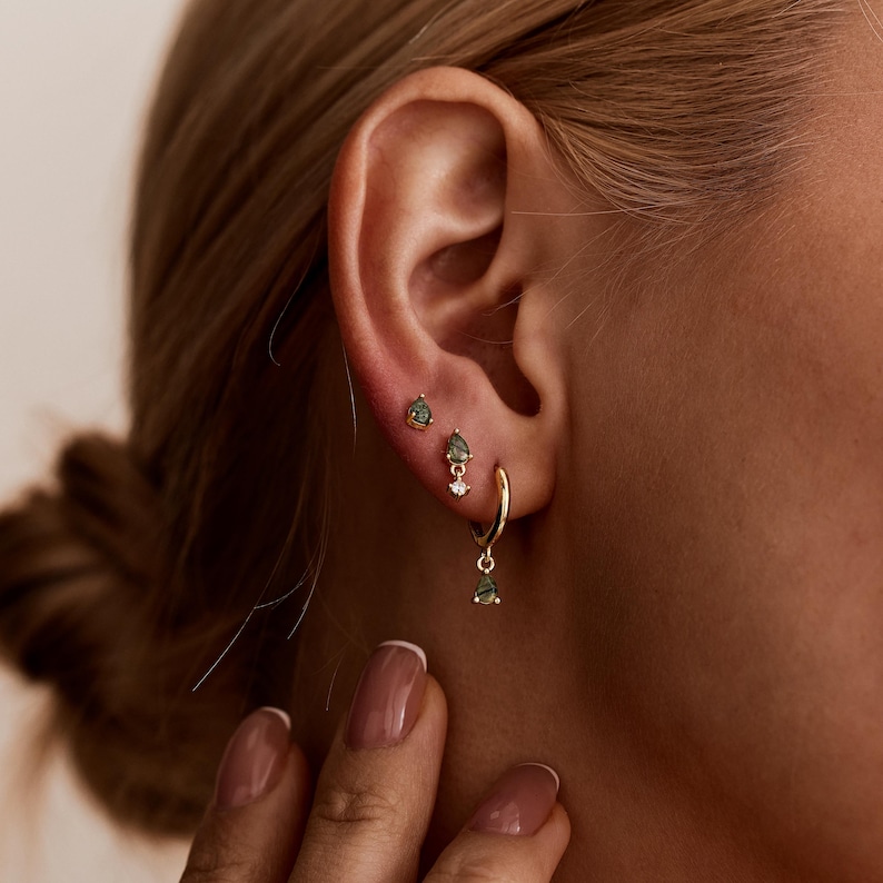 May include: A set of three gold earrings consisting of two small stud earrings with pear-shaped green stones and one gold hoop earring featuring a dangling pear-shaped green stone and a small clear round stone accent on the hoop.