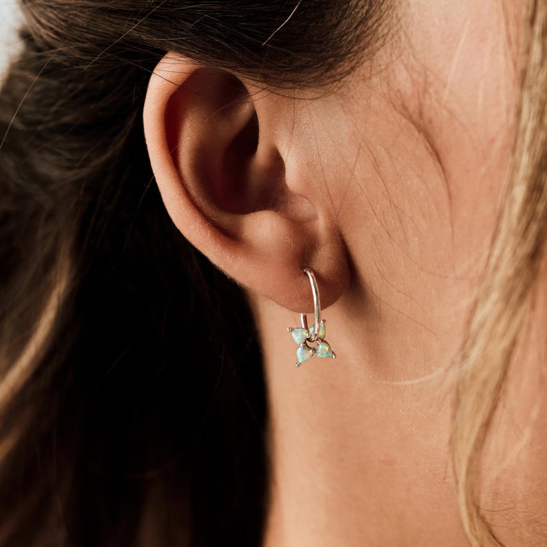 May include: A small silver hoop earring featuring a dangling charm composed of four teardrop-shaped iridescent opals arranged in a flower-like pattern. The earring is secured through the earlobe, showcasing the metallic silver band and the stones.