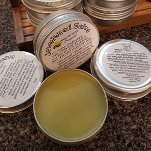 May include: A silver tin of Jewelweed Salve with a green herbal salve inside. The label reads "Jewelweed Salve" with the words "Hatchie River Soapworks" underneath. The label also lists the ingredients and benefits of the salve, including jewelweed, calendula, plantain, rosemary, and lavender essential oils. The salve is 100% natural and wildcrafted. It is 2 oz.