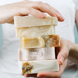 May include: A stack of six handmade soap bars in varying colors and textures. The soaps range from light beige to pale pink, with some featuring dried botanicals. The bars are held in a person's hands.