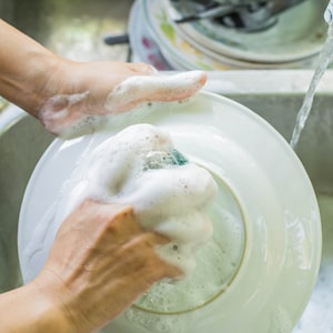 May include: A person's hands washing a white ceramic plate with soapy water and a sponge. The plate is in a sink with running water.