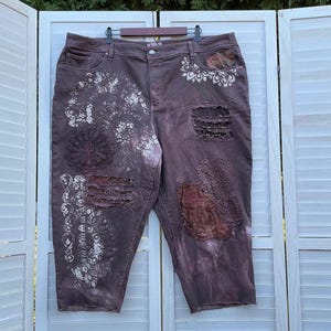 May include: Dark brown denim capri trousers featuring white floral lace-style stencilled patterns, distressed frayed sections, and dark brown fabric patches. The trousers include a button closure, belt loops, and five pockets with a faded wash finish.