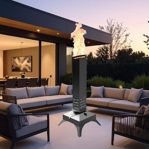 Op de afbeelding: Een hoge, zwarte buitenhaard met vlameffect. De haard heeft een vierkante basis met drie poten en een hoge, rechthoekige behuizing. De vlam is fel oranje en geel. De haard staat op een terras met tuinmeubelen.