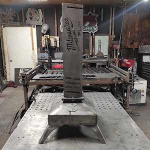 Puede incluir: Un brasero de metal con una chimenea alta y rectangular con el logotipo "Milwaukee FUEL" recortado. El brasero tiene una base cuadrada con patas anguladas y una puerta circular. El metal tiene un aspecto crudo y sin terminar.