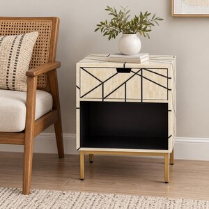 May include: A square bedside table featuring a cream-colored surface with a black geometric line pattern. The table includes one top drawer with a recessed handle and an open lower shelf. It is supported by four thin, gold-toned metal legs.