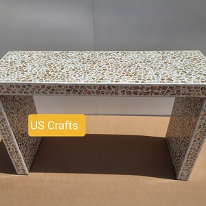 May include: A white console table with a top made of small, irregular pieces of iridescent shell in shades of white, brown, and gold. The table has a simple, rectangular design with a modern aesthetic. The text "US Crafts" is visible on the table.