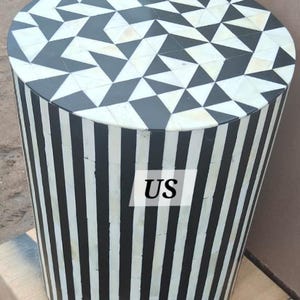 Puede incluir: Mesa auxiliar de incrustaciones de hueso en blanco y negro con un patrón geométrico en la parte superior y rayas verticales en el lateral. La mesa es redonda y tiene un diámetro de aproximadamente 46 cm.