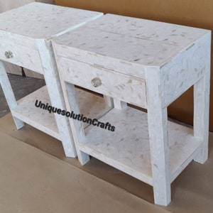 May include: Two white nightstands with a single drawer and a lower shelf. The nightstands are made of wood and have a white mother of pearl inlay.