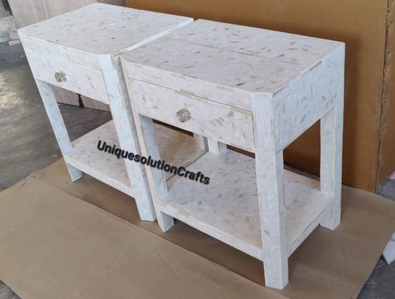 May include: Two white nightstands with a single drawer and a lower shelf. The nightstands are made of wood and have a white mother of pearl inlay.