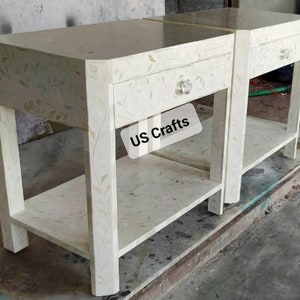 May include: Two white nightstands with a single drawer and a lower shelf. The nightstands are made of wood and have a floral design. The text "US Crafts" is visible on the front of one of the nightstands.