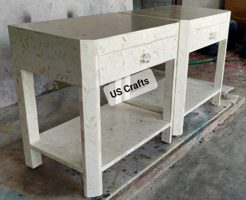 May include: Two white nightstands with a single drawer and a lower shelf. The nightstands are made of wood and have a floral design. The text "US Crafts" is visible on the front of one of the nightstands.