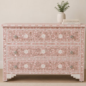 May include: A rectangular chest of drawers featuring a soft pink finish with intricate white floral and vine inlay patterns. The piece has three horizontal drawers, each equipped with two clear, faceted crystal-style knobs and stepped feet.