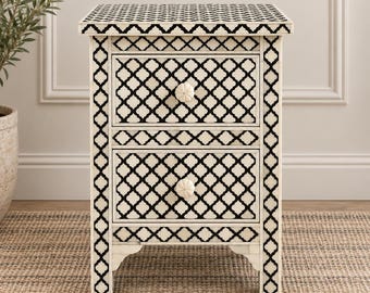 May include: A rectangular bedside table featuring two drawers with a black and white geometric lattice pattern. The piece has a flat top surface, four straight legs, and two round, off-white knobs centered on each drawer front.