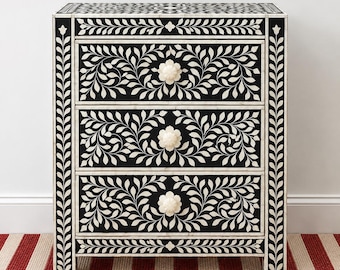 May include: A rectangular chest of drawers featuring three drawers with a black background and white floral vine inlay patterns. Each drawer has a round, off-white flower-shaped knob. The frame is decorated with a white leaf border design.