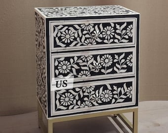 May include: A black and white bone inlay chest of drawers with a floral design. The chest has three drawers and sits on a gold metal stand. The chest is approximately 24 inches tall.