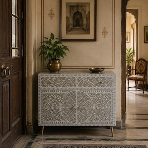 May include: A rectangular cabinet featuring a grey and white floral inlay pattern across its front surface. The piece includes two top drawers and two lower cabinet doors with small gold-toned knobs, supported by four slender, tapered gold-toned legs.