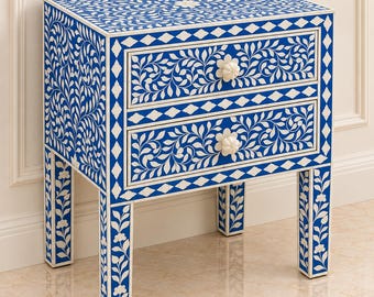May include: A rectangular bedside table featuring two drawers with white floral and vine patterns inlaid against a vibrant blue background. The piece includes four straight legs and two white flower-shaped knobs on the drawer fronts.